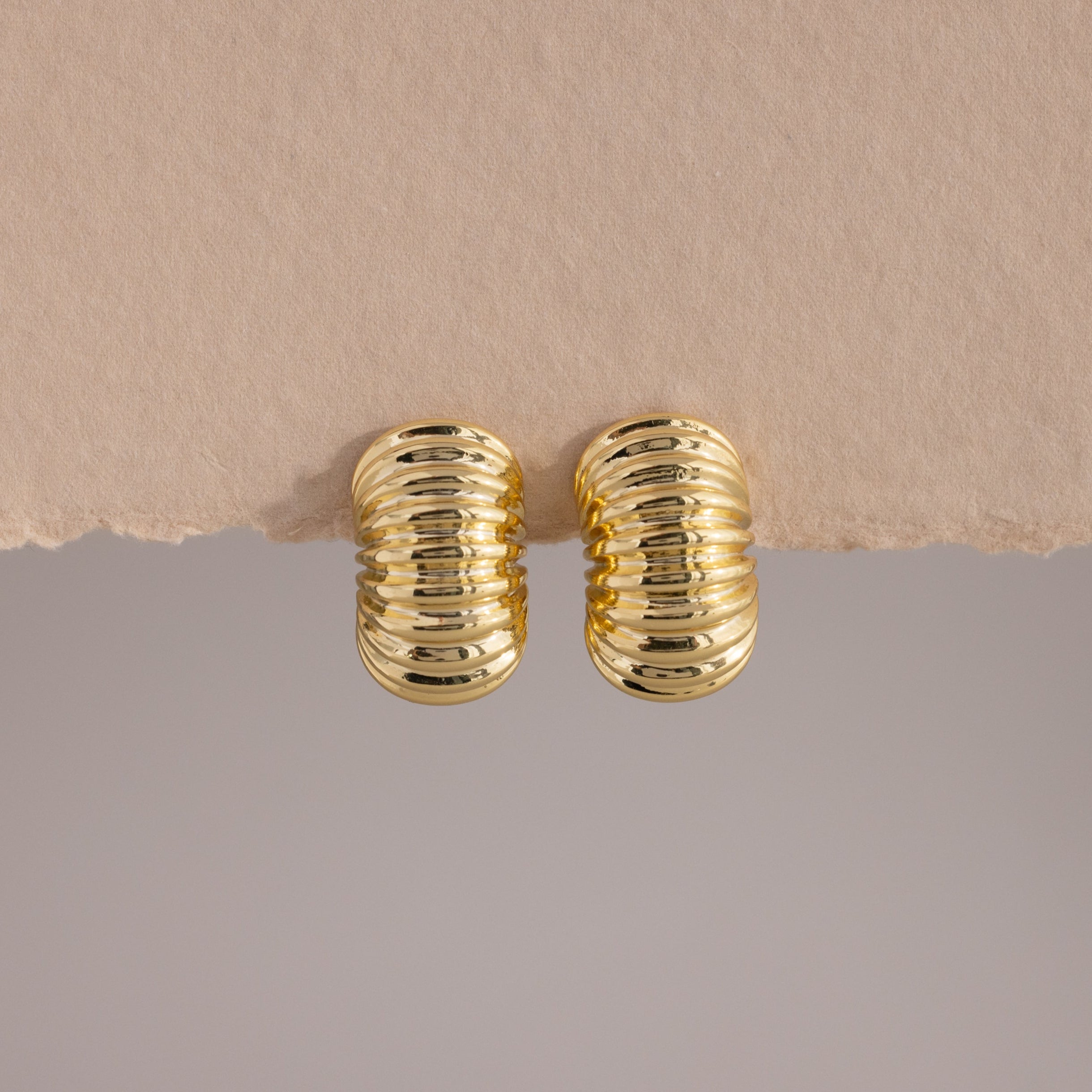 A pair of chunky, gold-toned ribbed hoop earrings displayed against a minimalist backdrop. The hoops feature a bold, domed silhouette with a high-polish finish and evenly spaced horizontal ridges that wrap around the surface, creating a sculptural, pillowy texture