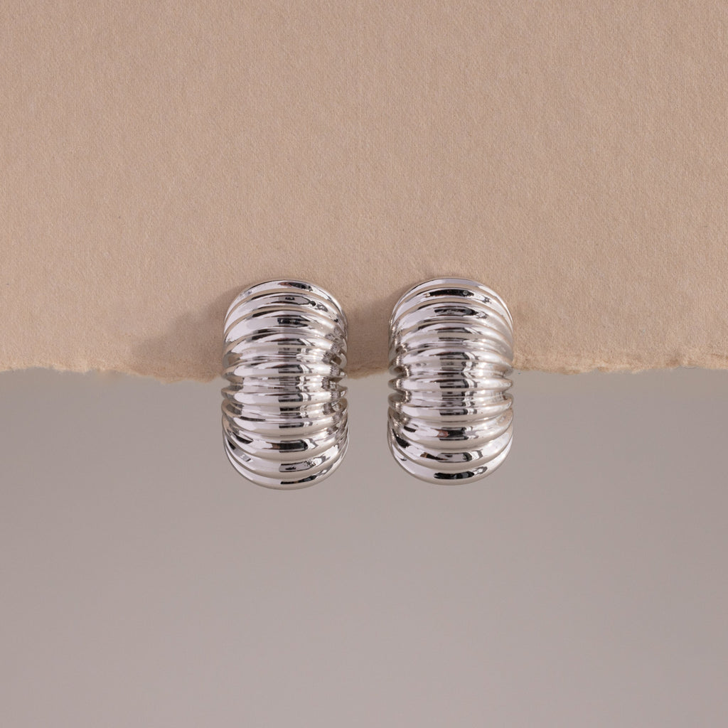 A pair of Chunky Ribbed Hoops in white gold, positioned upright and symmetrically on a lightly textured, torn-edge beige surface. The hoops have a bold, rounded silhouette with a series of prominent, high-polish ridges that create a corrugated effect