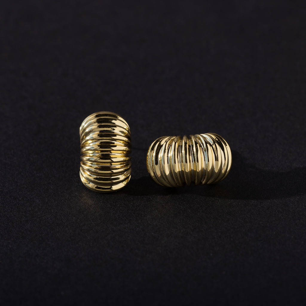 A pair of gold chunky ribbed hoop earrings placed on a dark background, with one earring standing upright and the other lying on its side