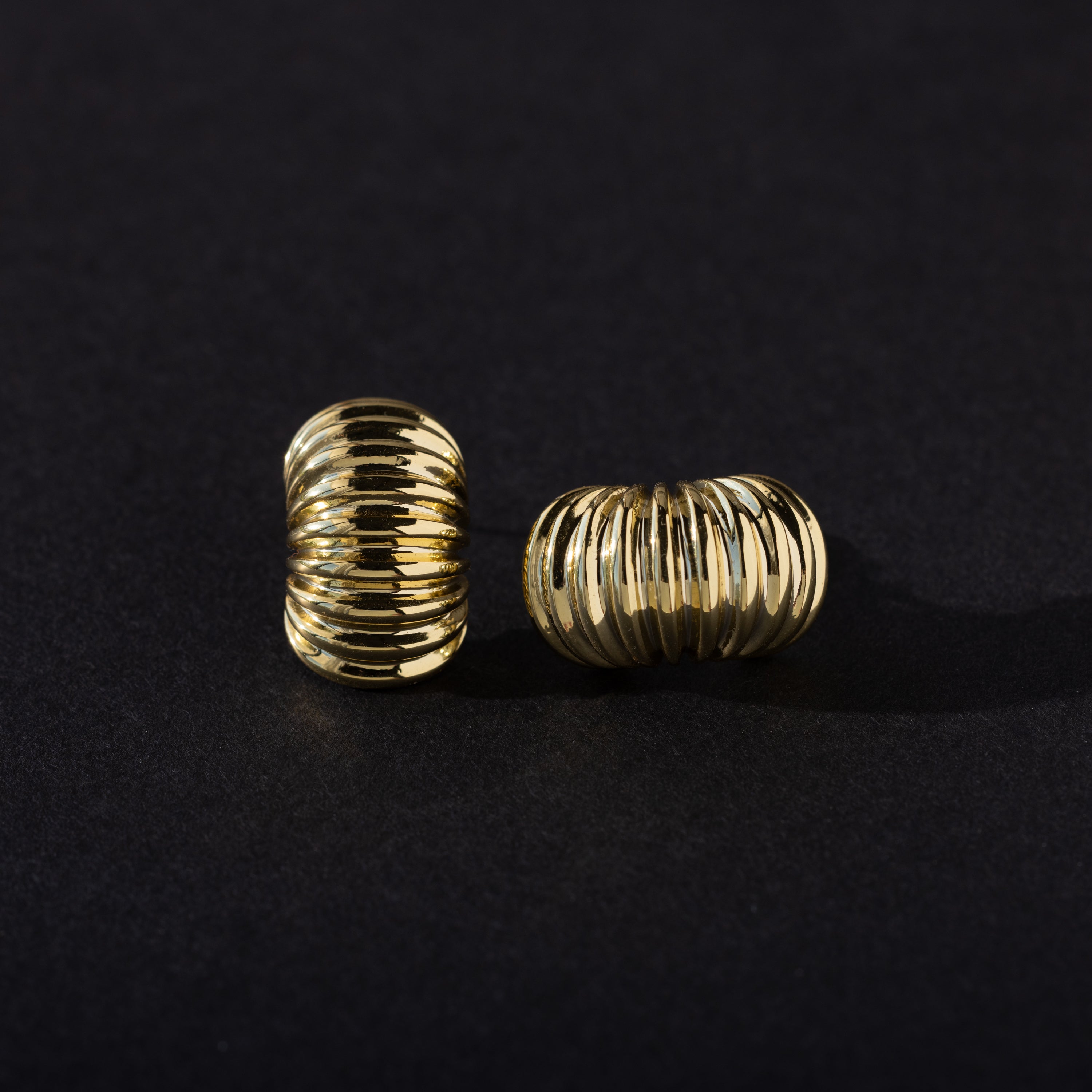 A pair of gold chunky ribbed hoop earrings placed on a dark background, with one earring standing upright and the other lying on its side