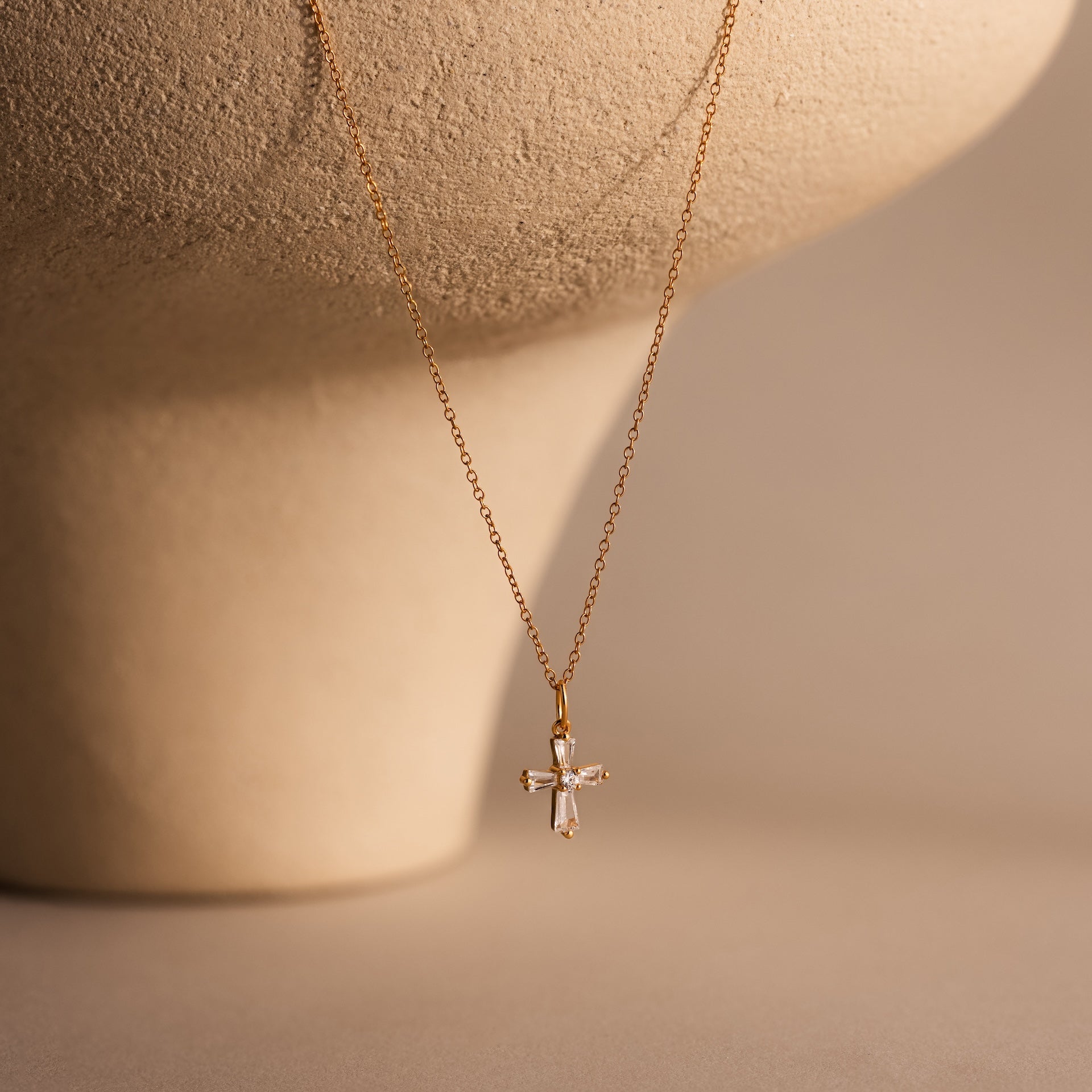 Art Deco Cross Necklace in 18K Gold, featuring a small jeweled pendant with baguette stones, displayed on a beige textured background—a timeless vintage jewelry piece.