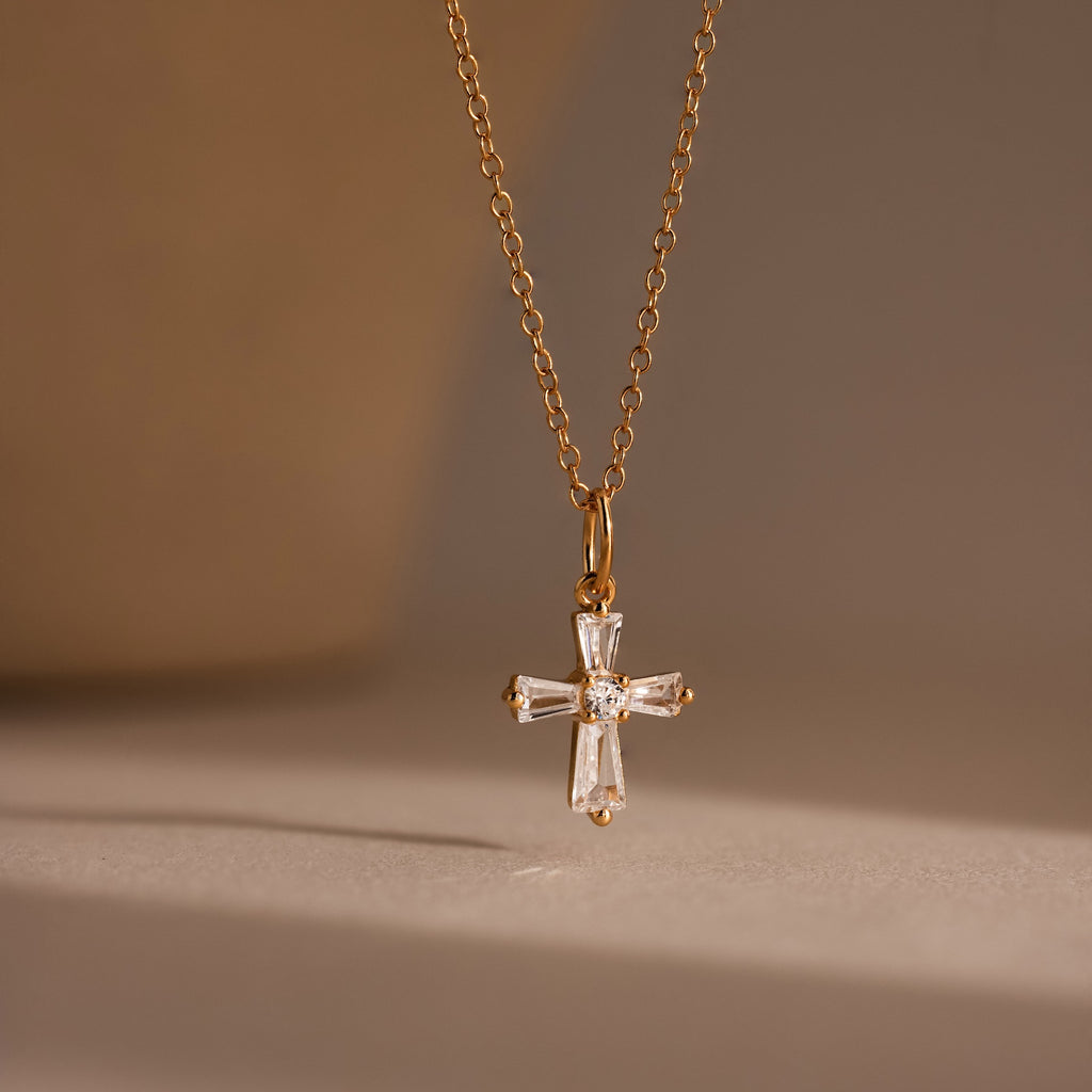 The Art Deco Cross Necklace in 18K Gold features a crystal cross pendant with baguette stones, showcased against a soft, neutral background.