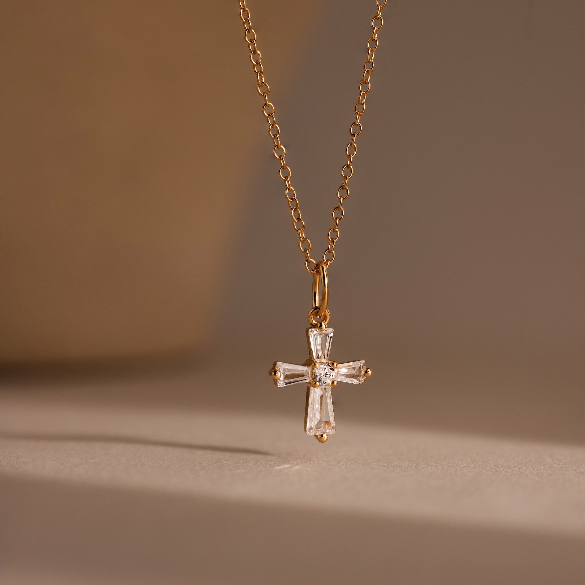 The Art Deco Cross Necklace in 18K Gold features a crystal cross pendant with baguette stones, showcased against a soft, neutral background.