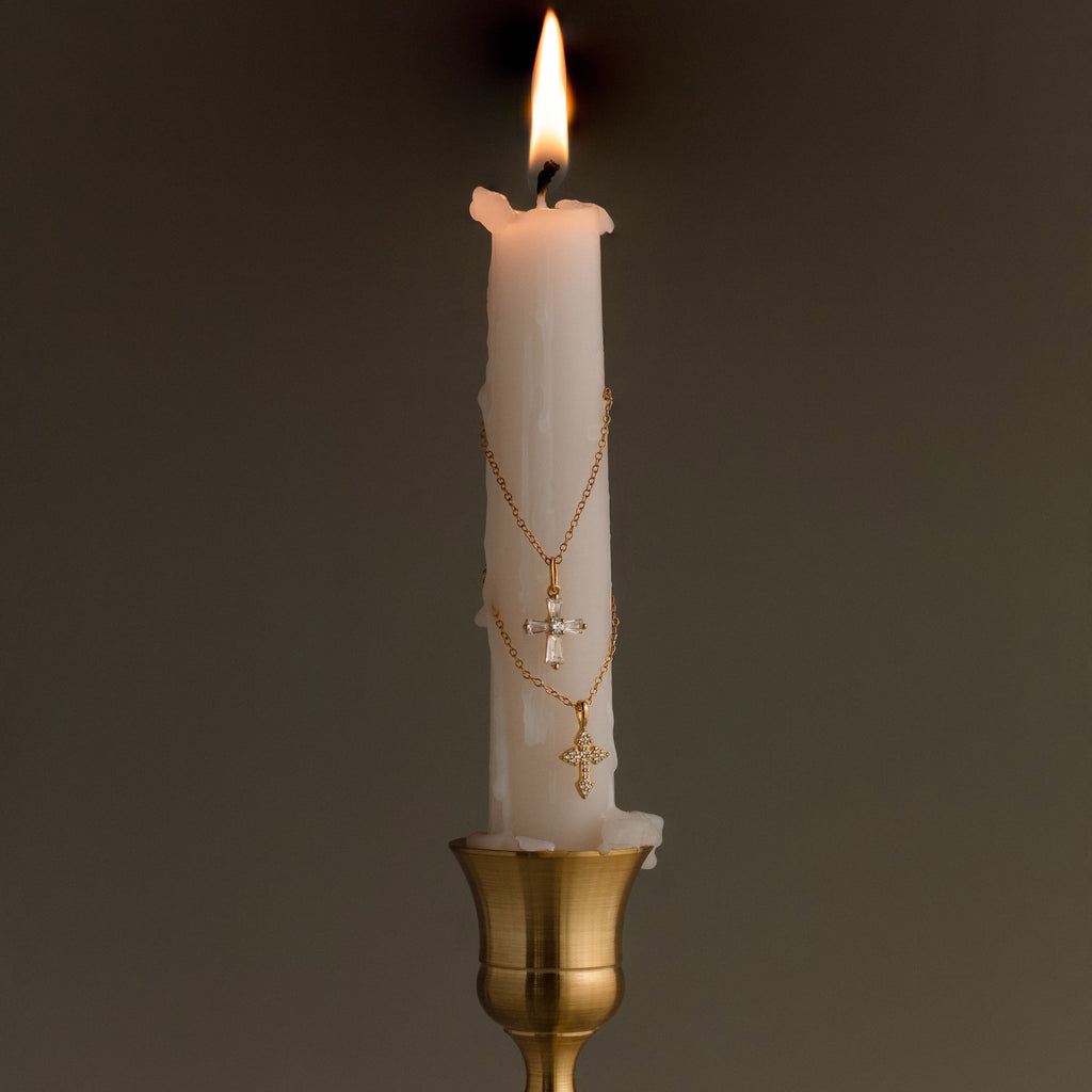 A lit white candle adorned with the Art Deco Cross Necklace and gold chains draped around it sits in a brass holder against a plain background.