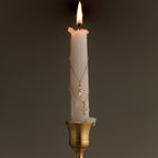 A lit white candle adorned with the Art Deco Cross Necklace and gold chains draped around it sits in a brass holder against a plain background.