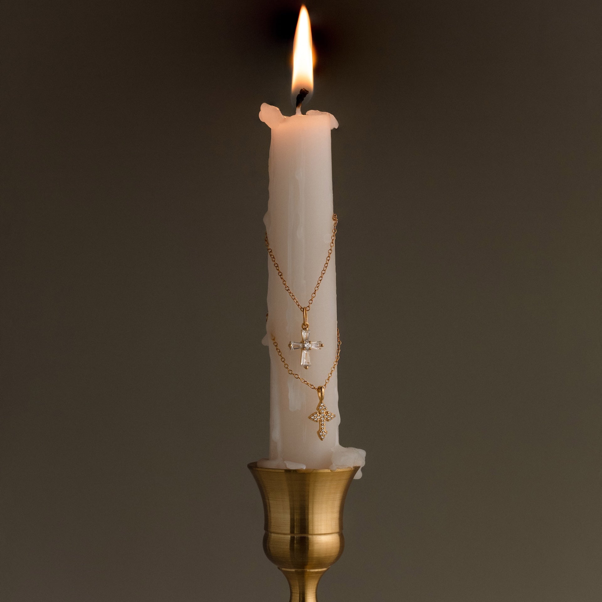 A lit white candle adorned with the Art Deco Cross Necklace and gold chains draped around it sits in a brass holder against a plain background.