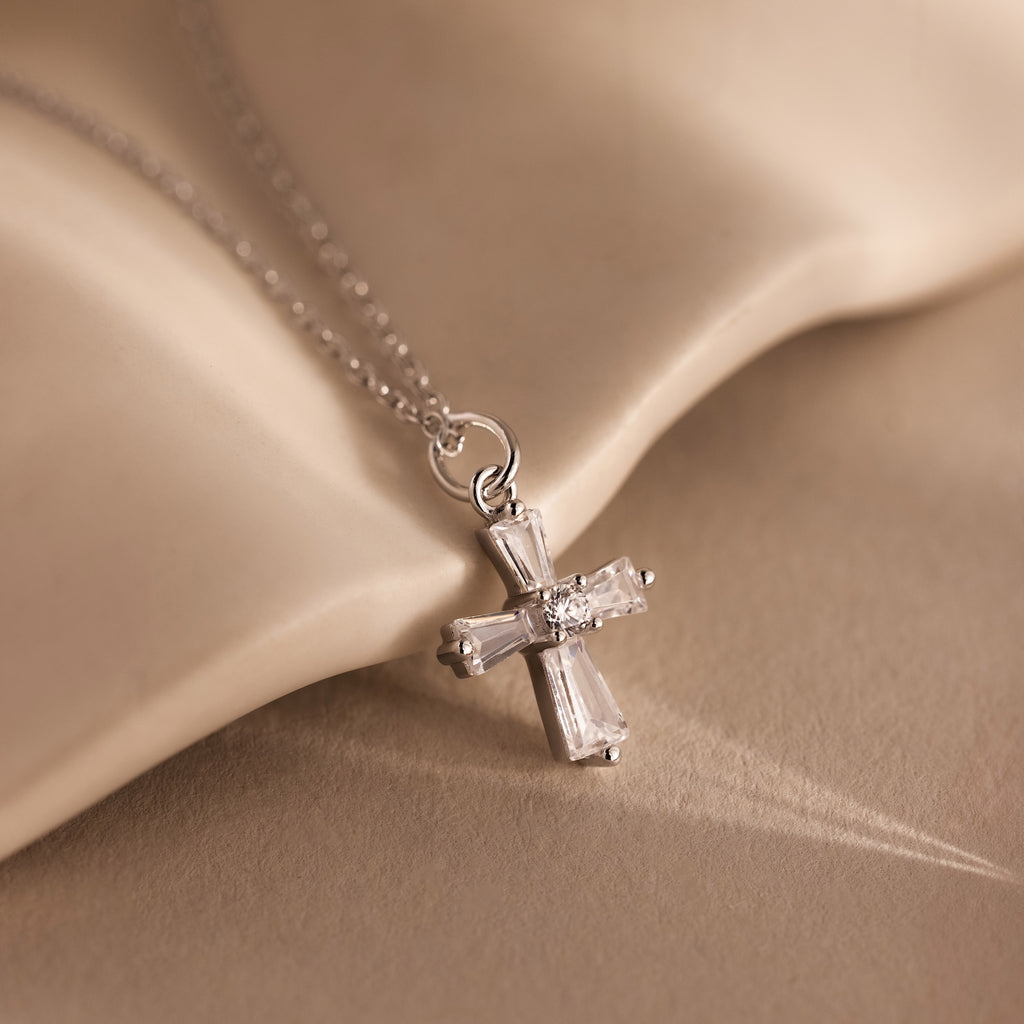 The Art Deco Cross Necklace in Silver features a delicate silver cross pendant with a small diamond, beautifully displayed against a soft beige fabric background.