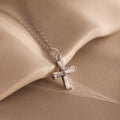 The Art Deco Cross Necklace in Silver features a delicate silver cross pendant with a small diamond, beautifully displayed against a soft beige fabric background.