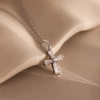 The Art Deco Cross Necklace in Silver features a delicate silver cross pendant with a small diamond, beautifully displayed against a soft beige fabric background.