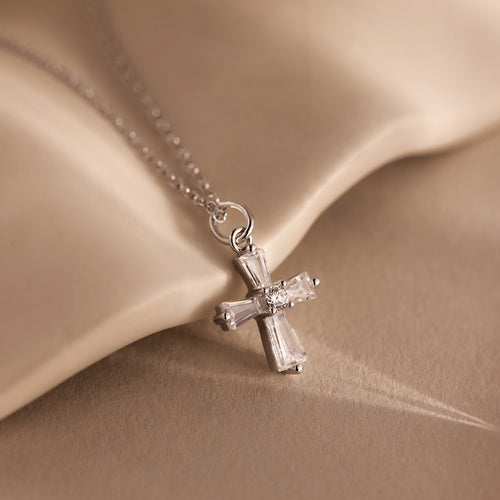 The Art Deco Cross Necklace in Silver features a delicate silver cross pendant with a small diamond, beautifully displayed against a soft beige fabric background.