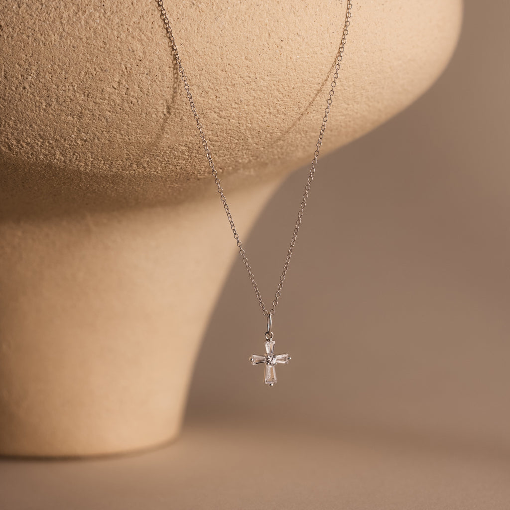 Art Deco Cross Necklace in Silver with baguette stones hangs on a delicate chain, shown against a beige textured background for vintage jewelry charm.
