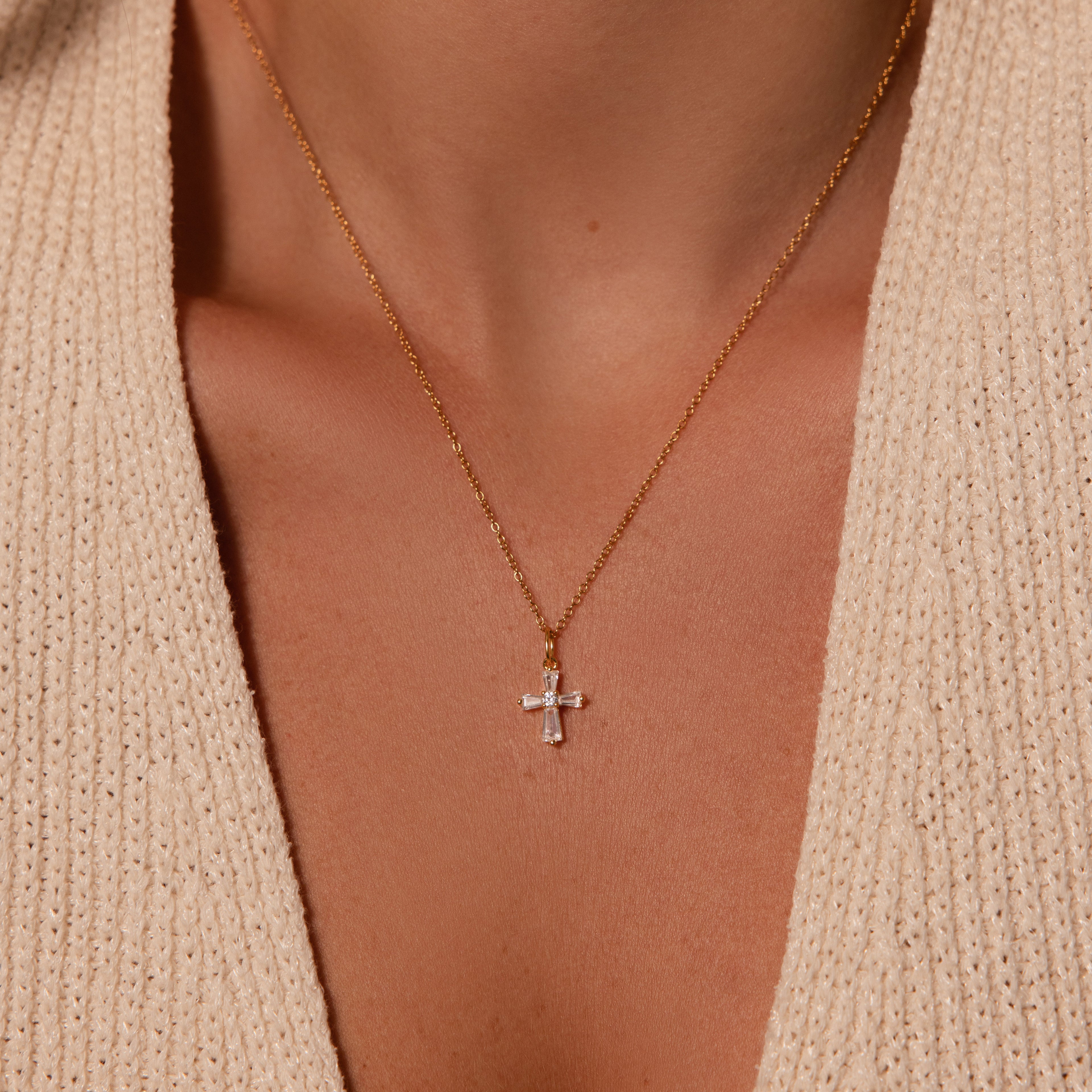 The Art Deco Cross Necklace features a dainty baguette diamond cross pendant on a gold chain, styled over a beige knit sweater.