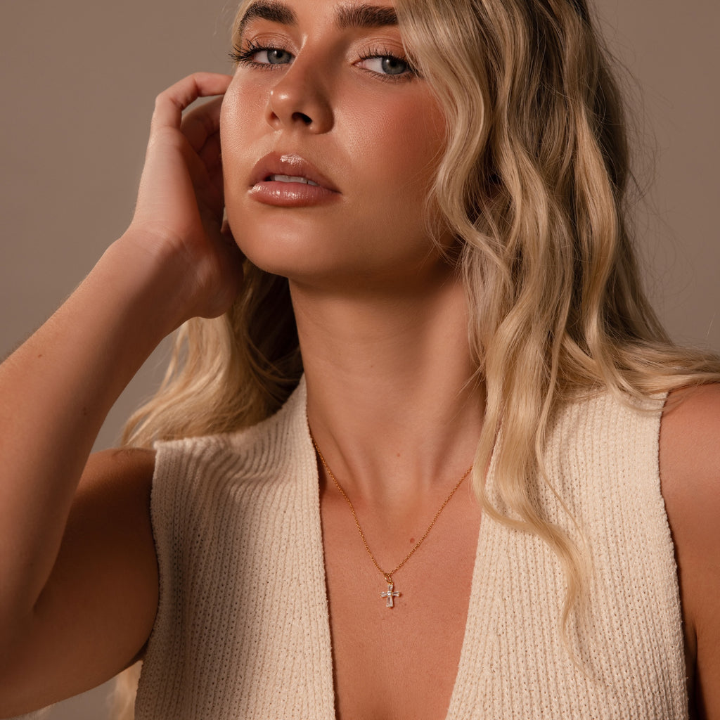 A woman with long blonde hair wears a cream knit top and the Art Deco Cross Necklace in 18K Gold, adding a touch of vintage jewelry elegance to her look.