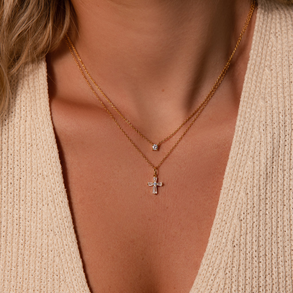 Close-up of a woman wearing two gold necklaces over a beige top, including the Art Deco Cross Necklace in 18K Gold featuring baguette stones and a small pendant necklace.