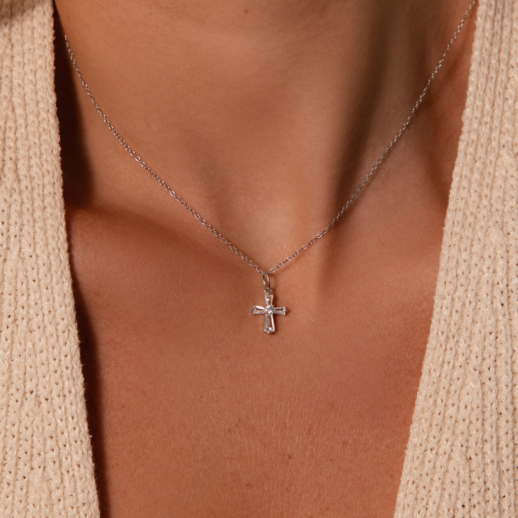 The Art Deco Cross Necklace in Silver, featuring baguette stones, is stylishly worn around the neck and pairs elegantly with a beige textured top.