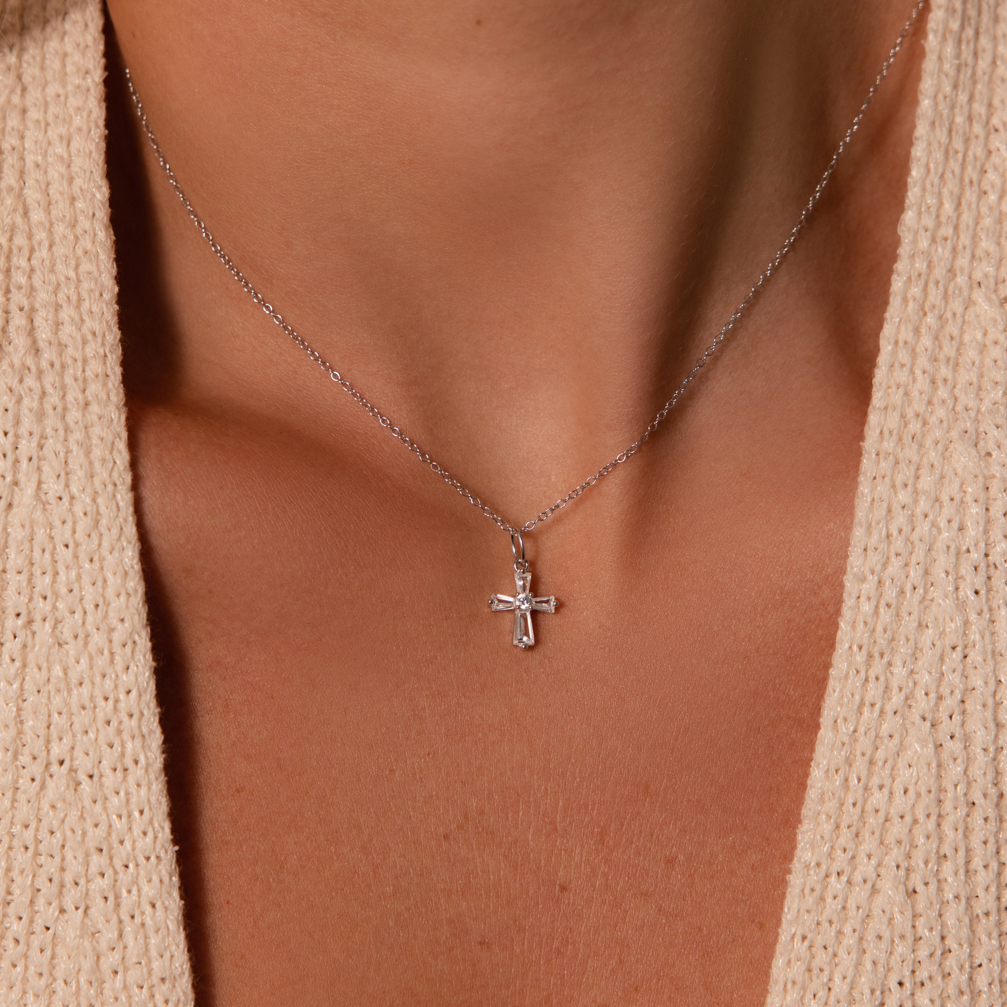 The Art Deco Cross Necklace in Silver, featuring baguette stones, is stylishly worn around the neck and pairs elegantly with a beige textured top.