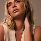 A woman with wavy blonde hair gazes upward, touching her neck, wearing a sleeveless cream top and the Art Deco Cross Necklace in Silver adorned with sparkling baguette stones.
