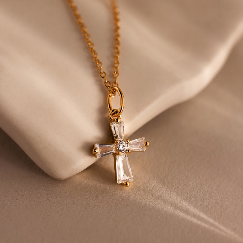 The Art Deco Cross Necklace in 18K Gold, adorned with crystal and baguette stones, is beautifully displayed on a soft beige surface.