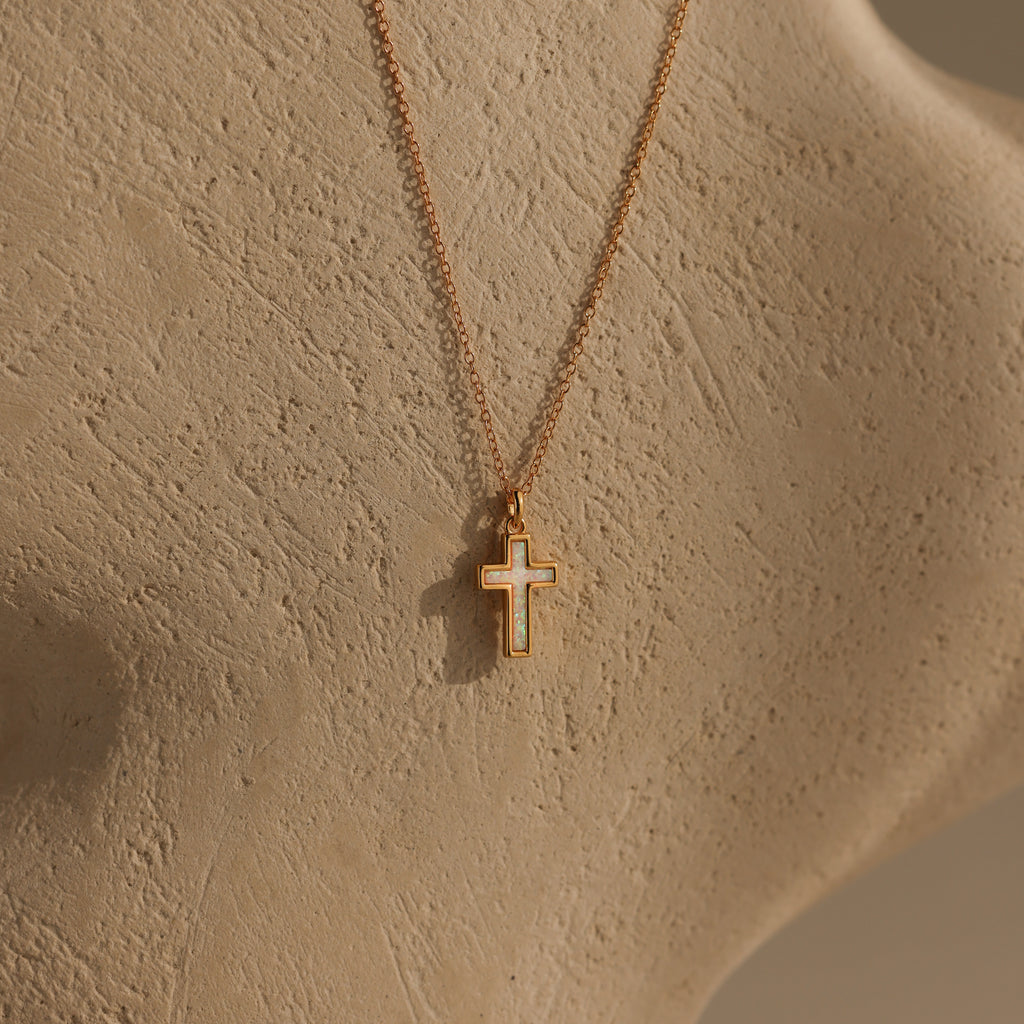 Mother of Pearl Cross Necklace in 18K Gold featuring an iridescent inlay, displayed on a textured beige surface and softly lit to highlight its minimalistic and timeless appeal.