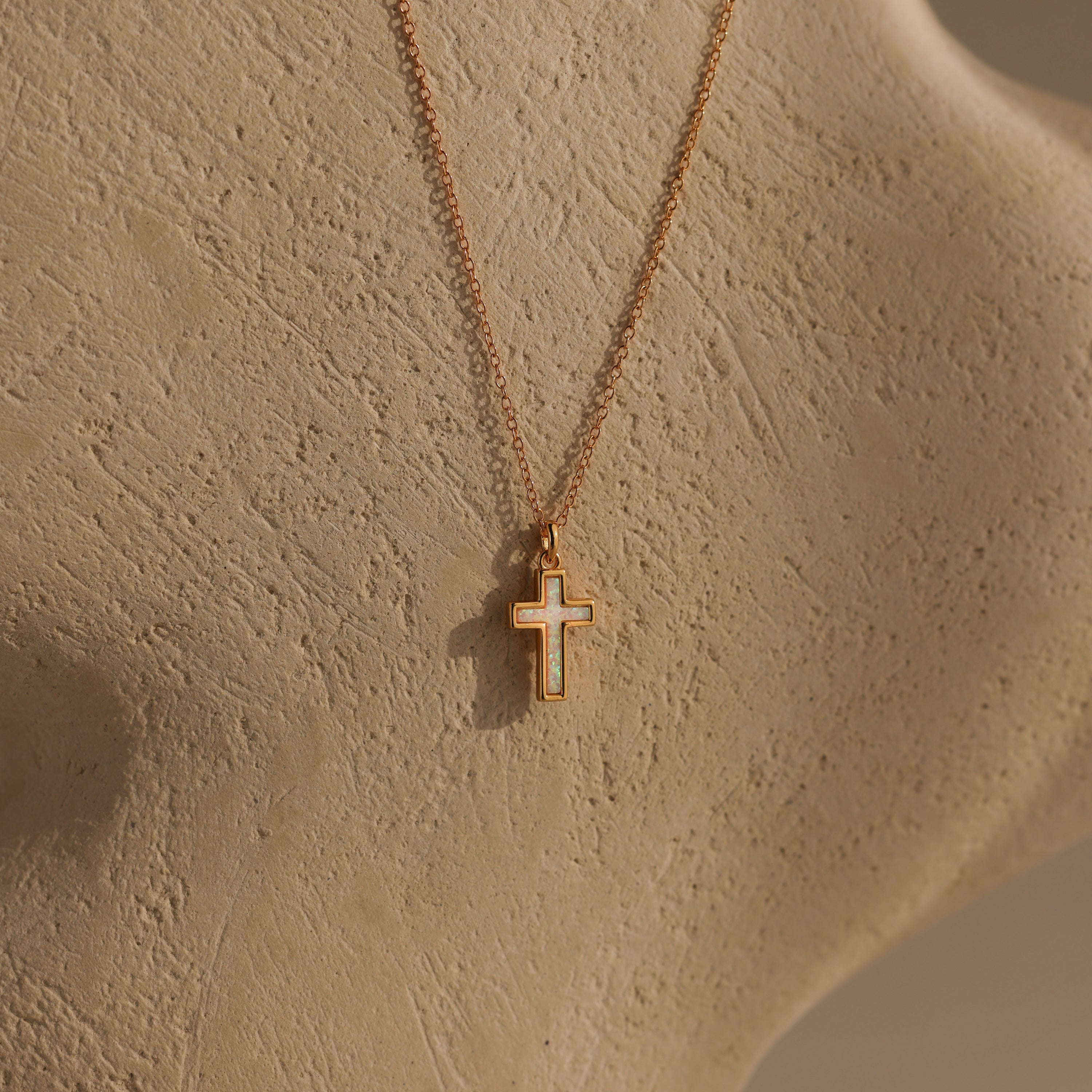 Mother of Pearl Cross Necklace in 18K Gold featuring an iridescent inlay, displayed on a textured beige surface and softly lit to highlight its minimalistic and timeless appeal.