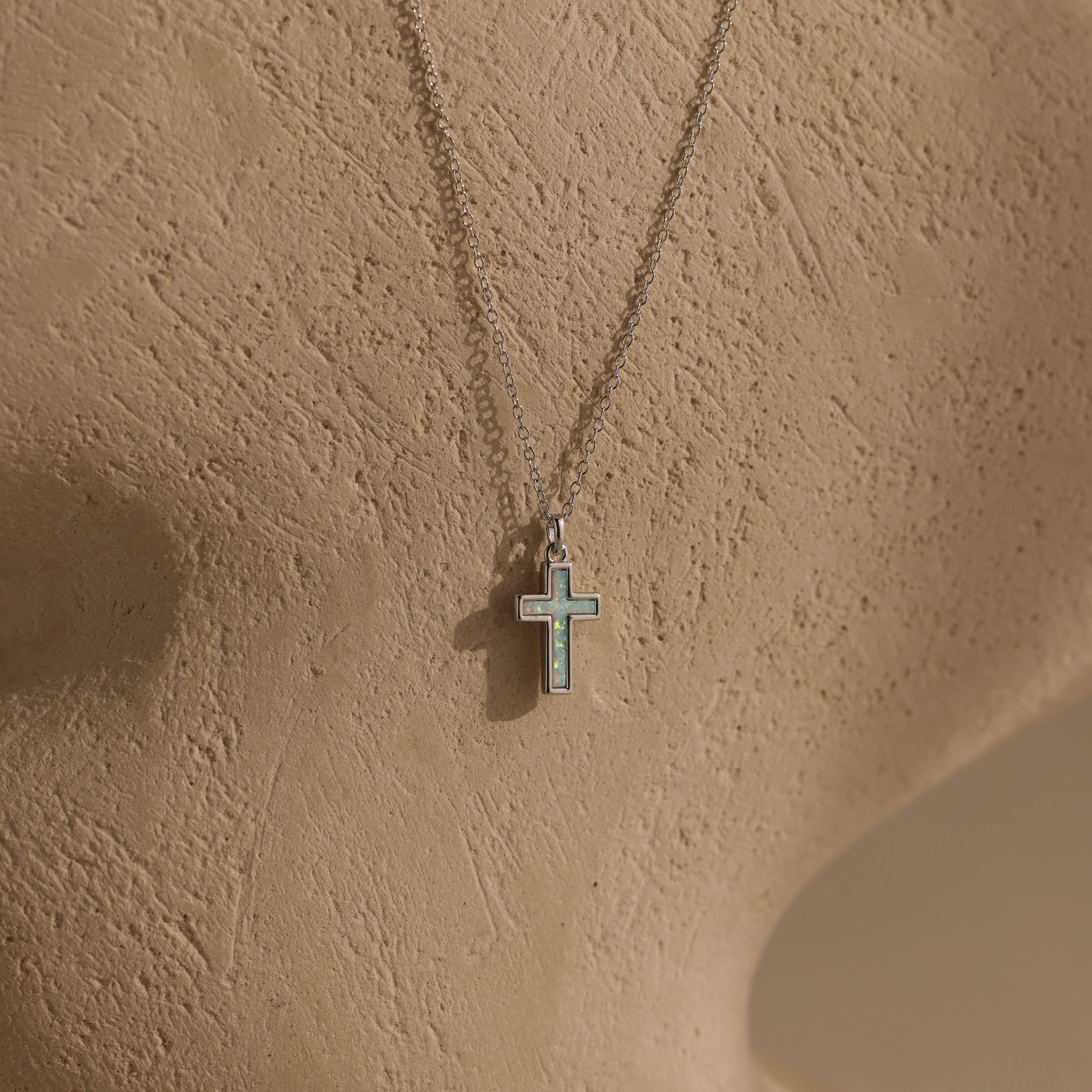 The Mother of Pearl Cross Necklace features a small iridescent pendant on a silver chain, displayed on a textured beige mannequin bust, highlighting timeless jewelry style.