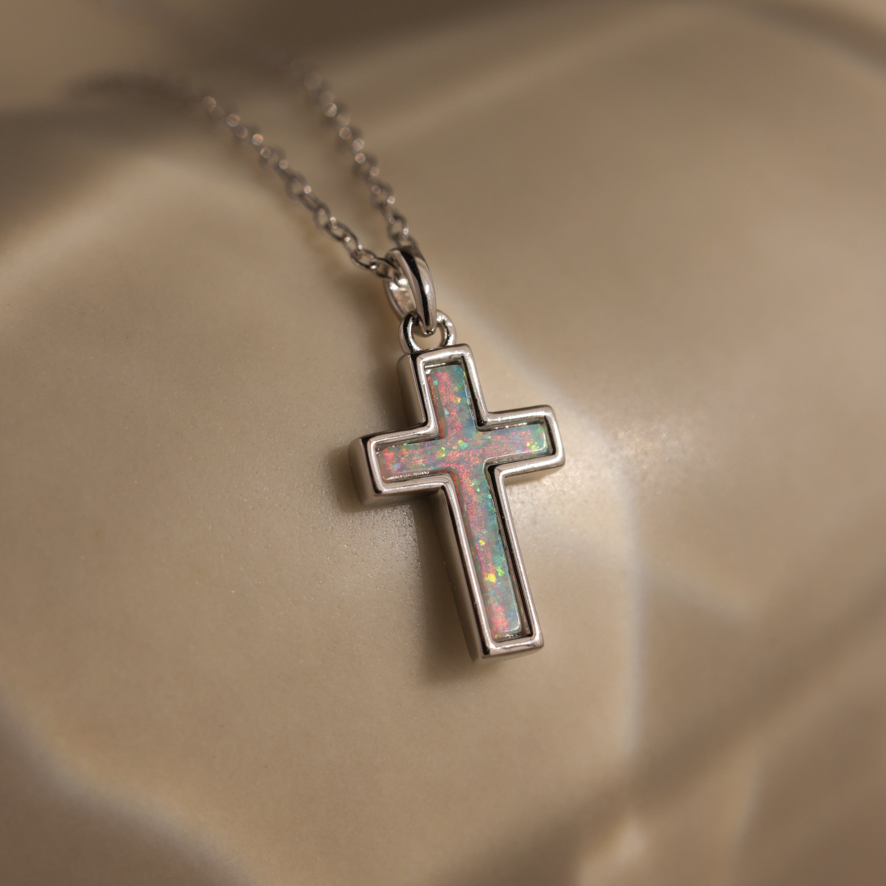 The Mother of Pearl Cross Necklace in Silver, featuring an iridescent inlay, is displayed on a smooth beige surface—a timeless jewelry piece that elevates any outfit.