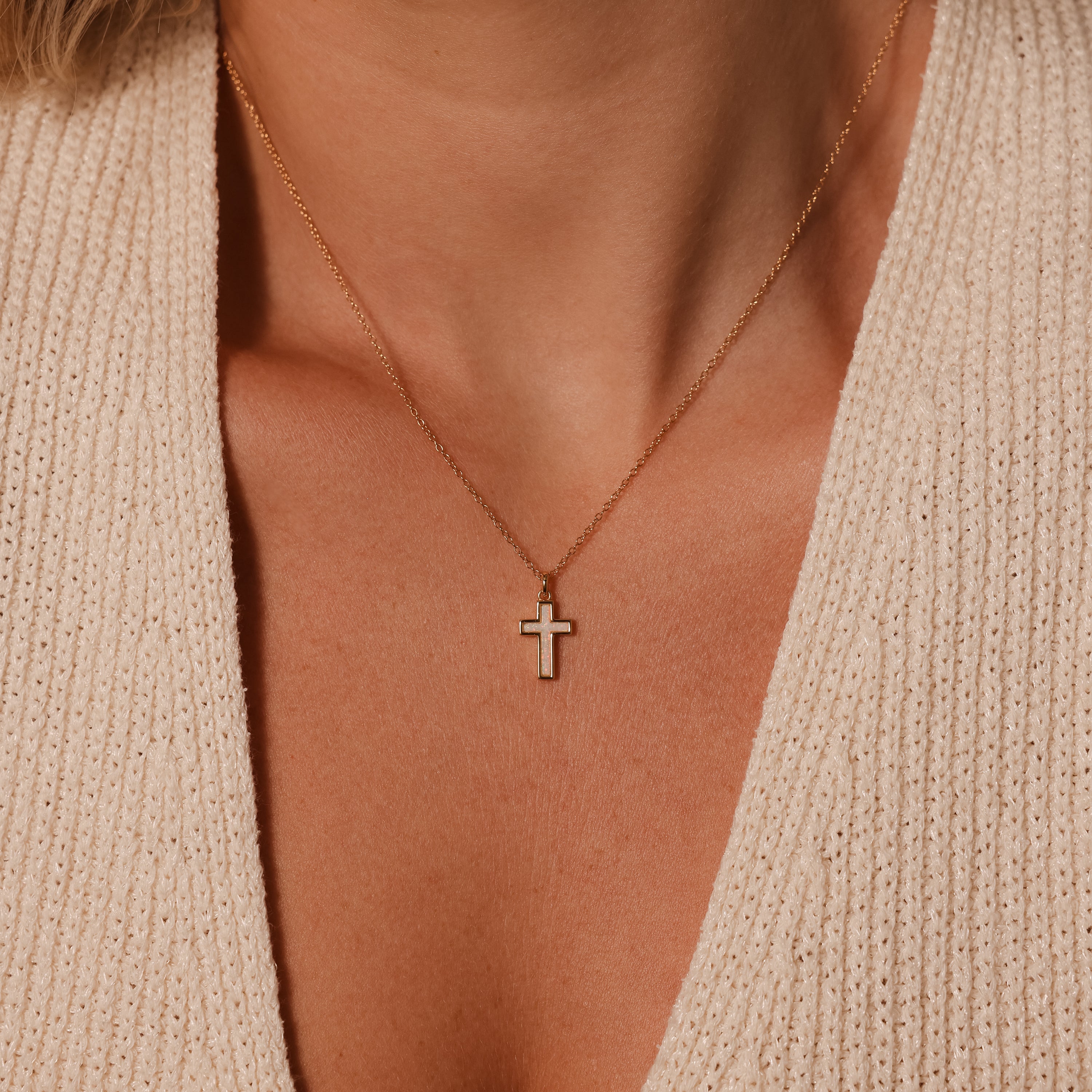 A woman in a beige knit deep V-neck top wears the Mother of Pearl Cross Necklace, featuring a gold chain and iridescent inlay, adding timeless elegance to her look.