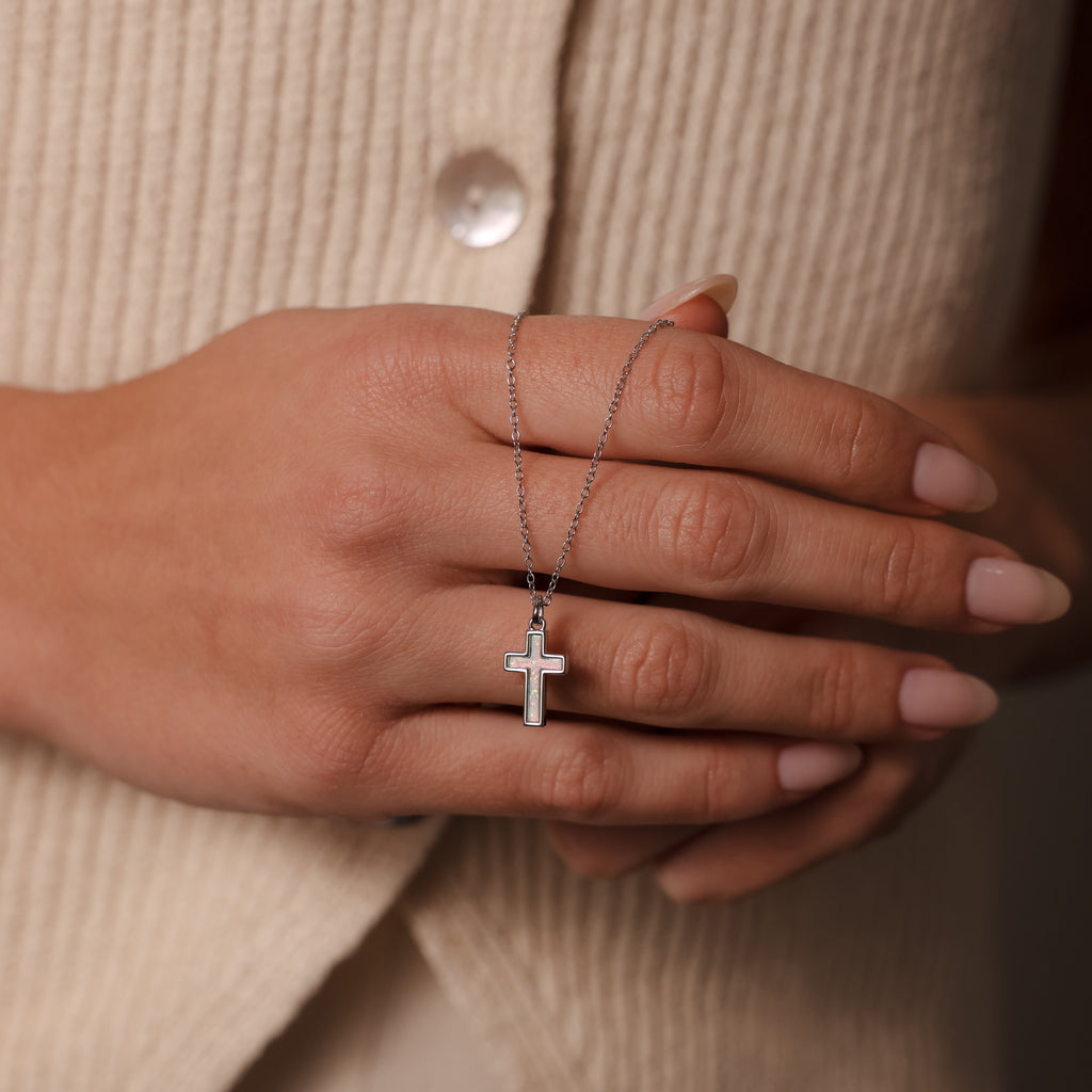 A manicured hand holds the Mother of Pearl Cross Necklace in Silver, featuring an iridescent inlay, set against a cream ribbed buttoned top—a timeless jewelry piece.