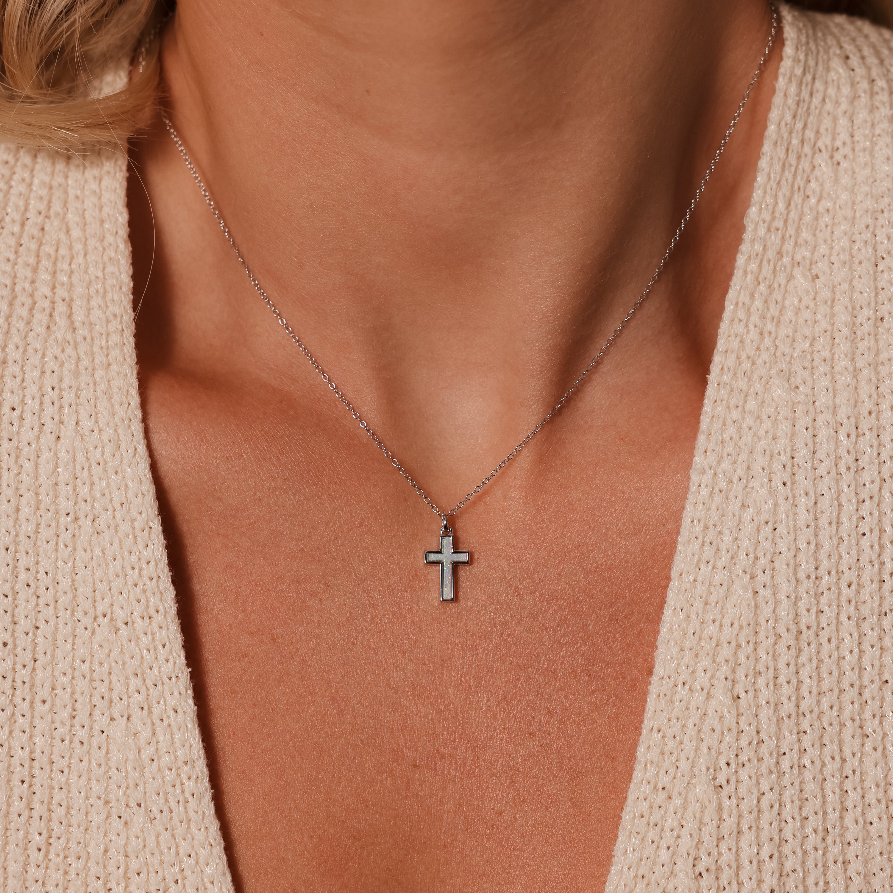 The Mother of Pearl Cross Necklace in Silver rests on her neck, its iridescent inlay glowing softly against a beige knit top with a deep neckline—a timeless piece of jewelry.