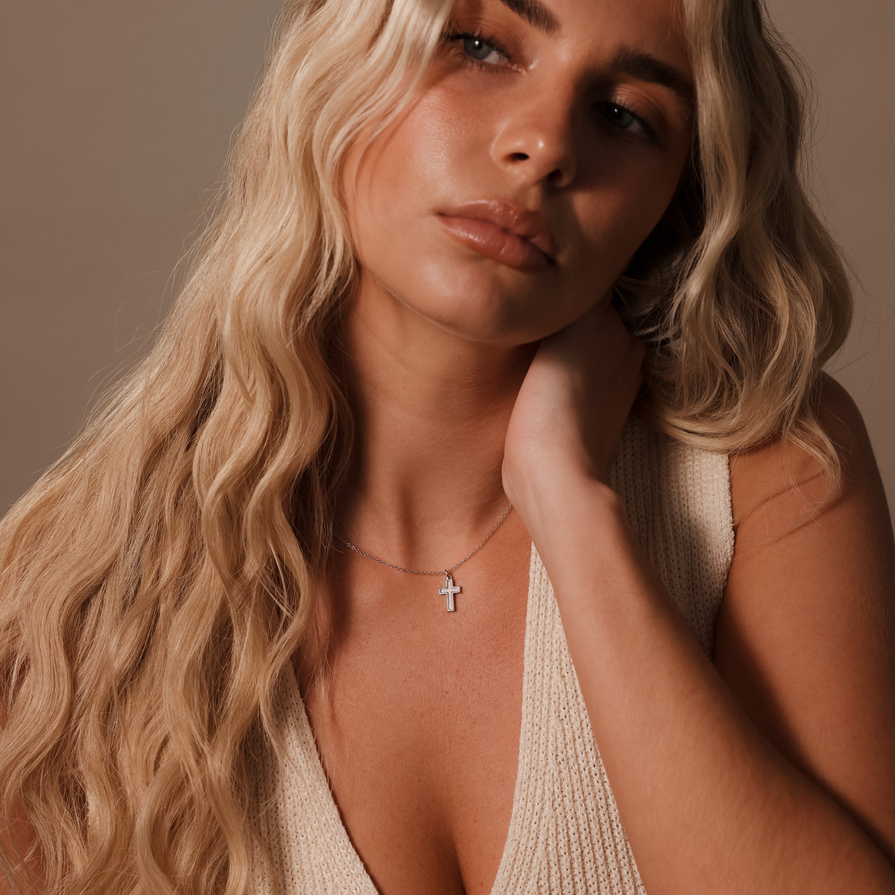 A woman with long blonde hair and a cream knit top wears the Mother of Pearl Cross Necklace, the timeless accessory gleaming as she looks to the side.