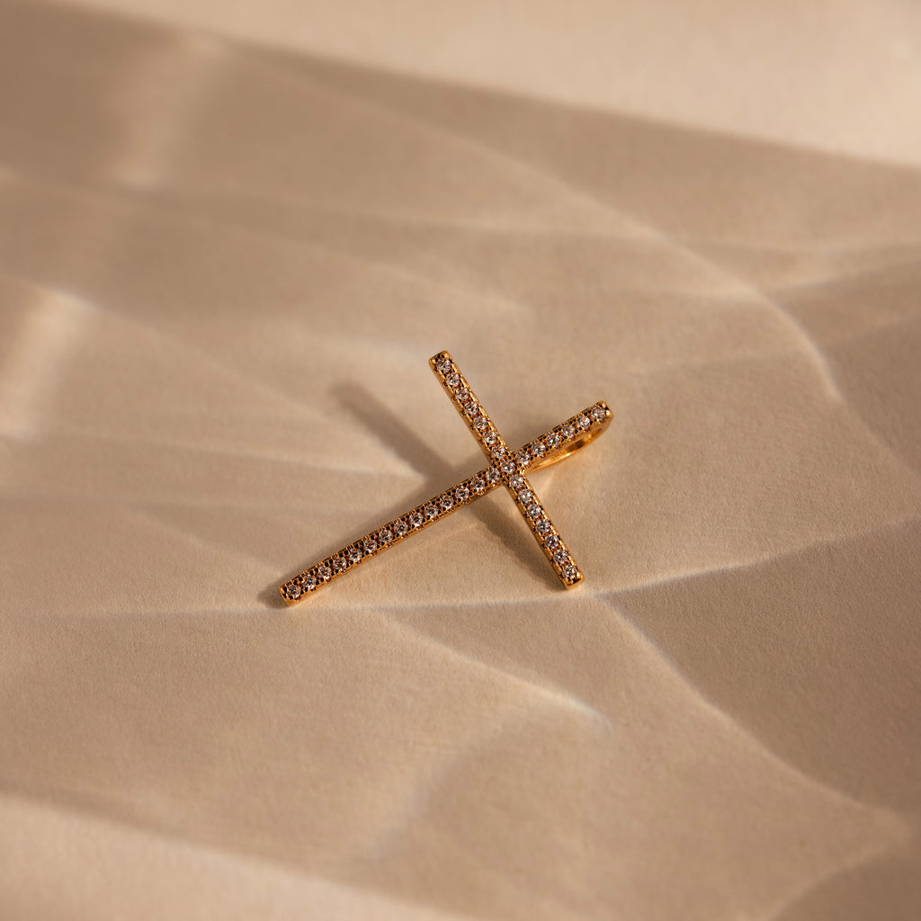 The Skinny Pave Cross Charm in 18K Gold with small diamonds rests on a light beige textured surface, perfect for fans of elegant faith jewelry.