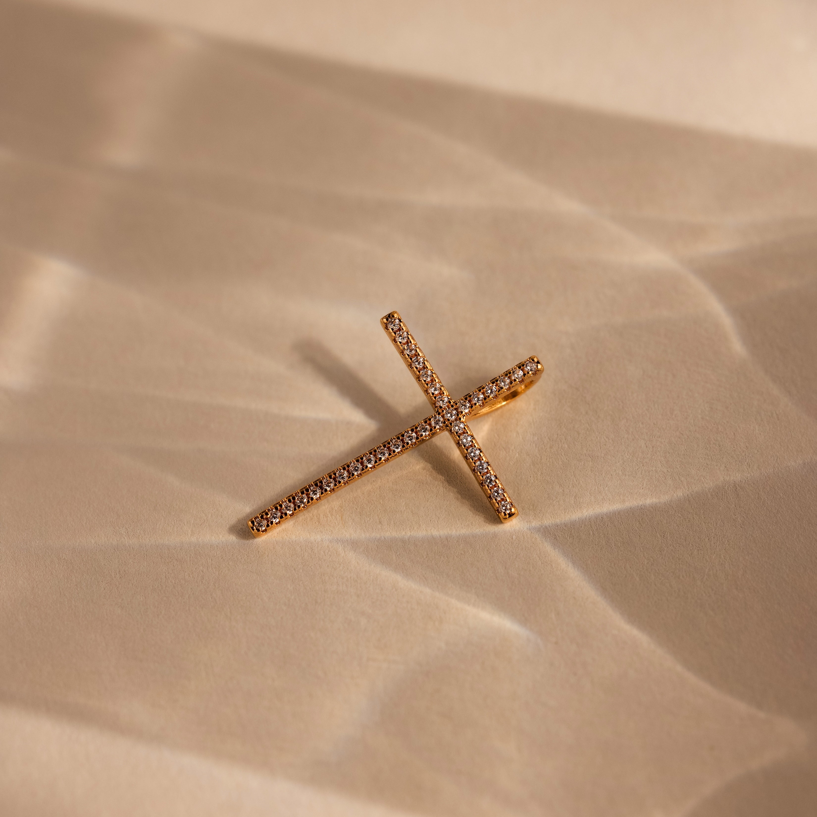 The Skinny Pave Cross Charm in 18K Gold with small diamonds rests on a light beige textured surface, perfect for fans of elegant faith jewelry.