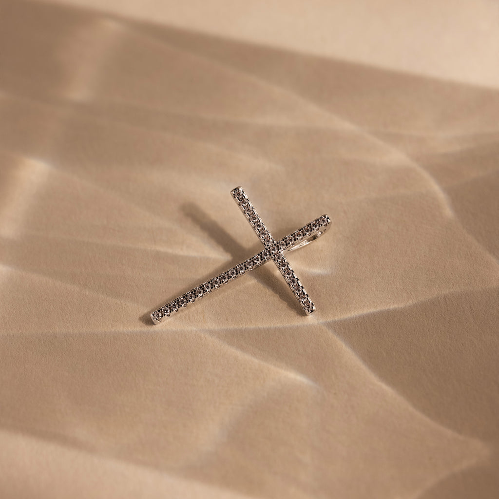 The Skinny Pave Cross Charm in Silver, sparkling with diamonds, lies on textured beige sand highlighted by soft shadows.