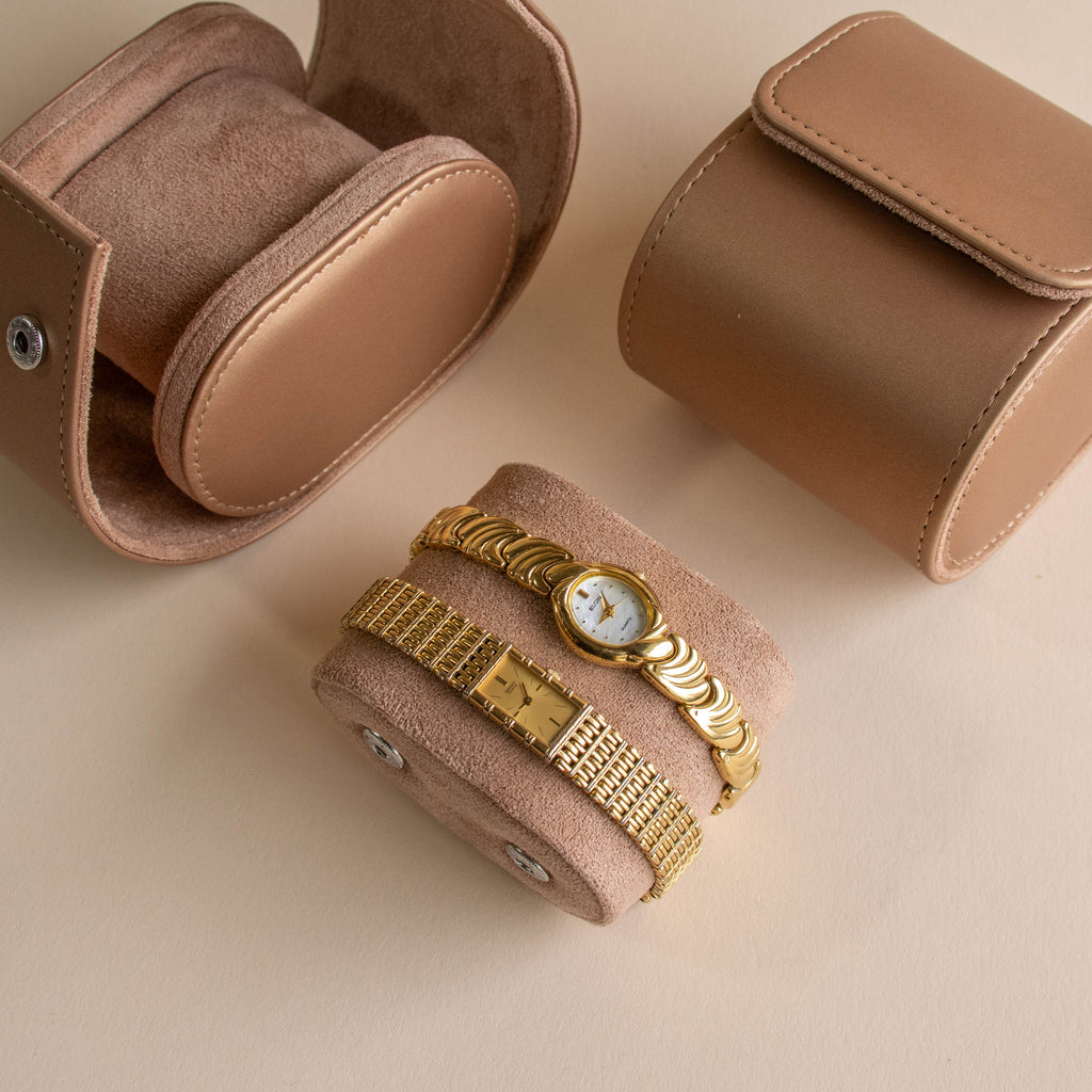 The Petite Oval Link Timepiece and another gold-tone watch are displayed on a suede roll, with brown watch cases resting on a beige surface.