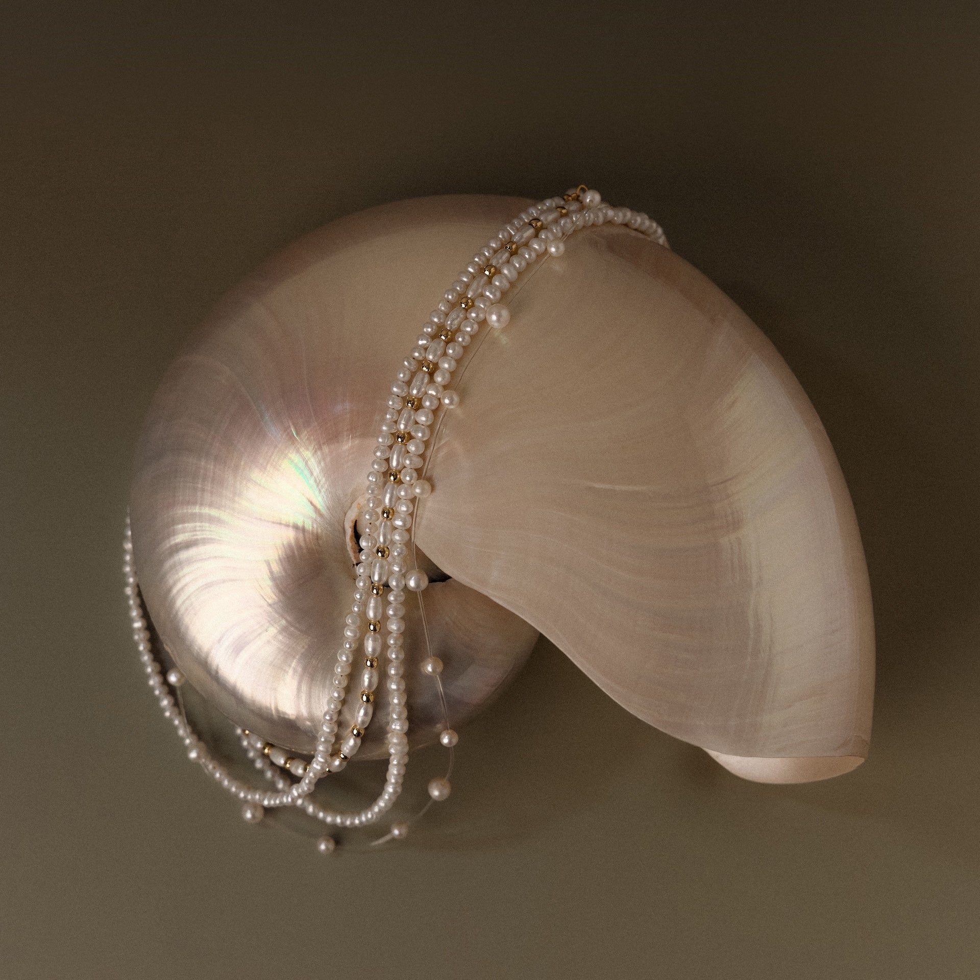 The Summer Pearl Beaded Necklace in 18K Gold is draped elegantly over a shiny spiral seashell on a simple neutral background.