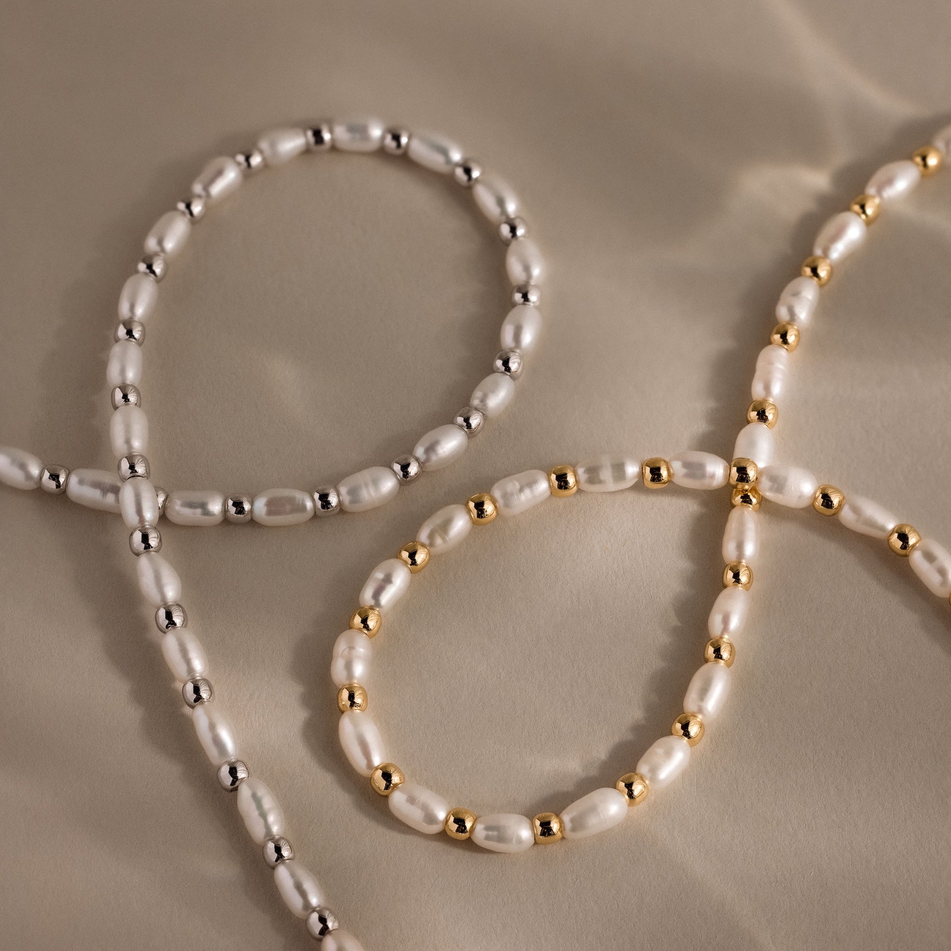 Two Beaded Pearl Necklaces, one featuring polished silver beads and the other gold accents, are arranged on a soft beige surface.