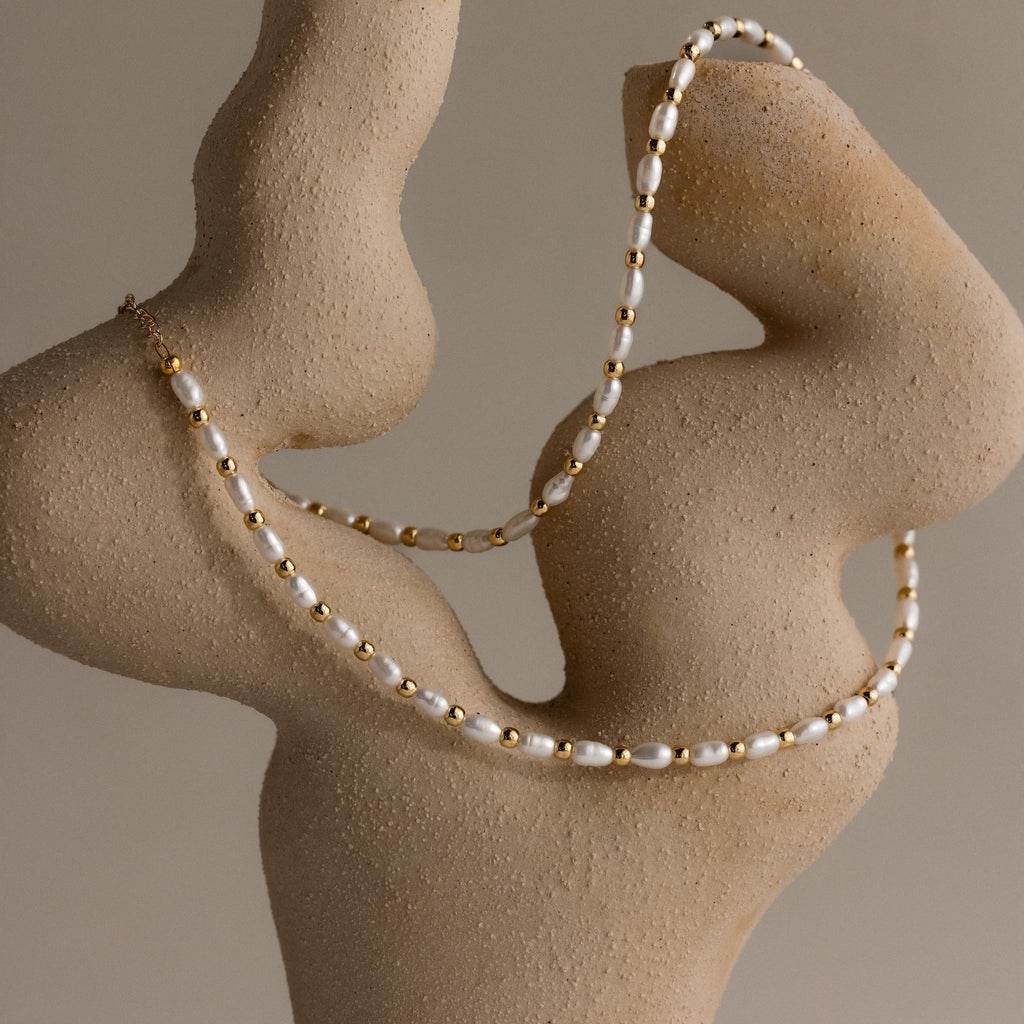 The Beaded Pearl Necklace in 18K Gold, featuring delicate polished beads and freshwater pearls, is elegantly draped over a textured, abstract beige sculpture.
