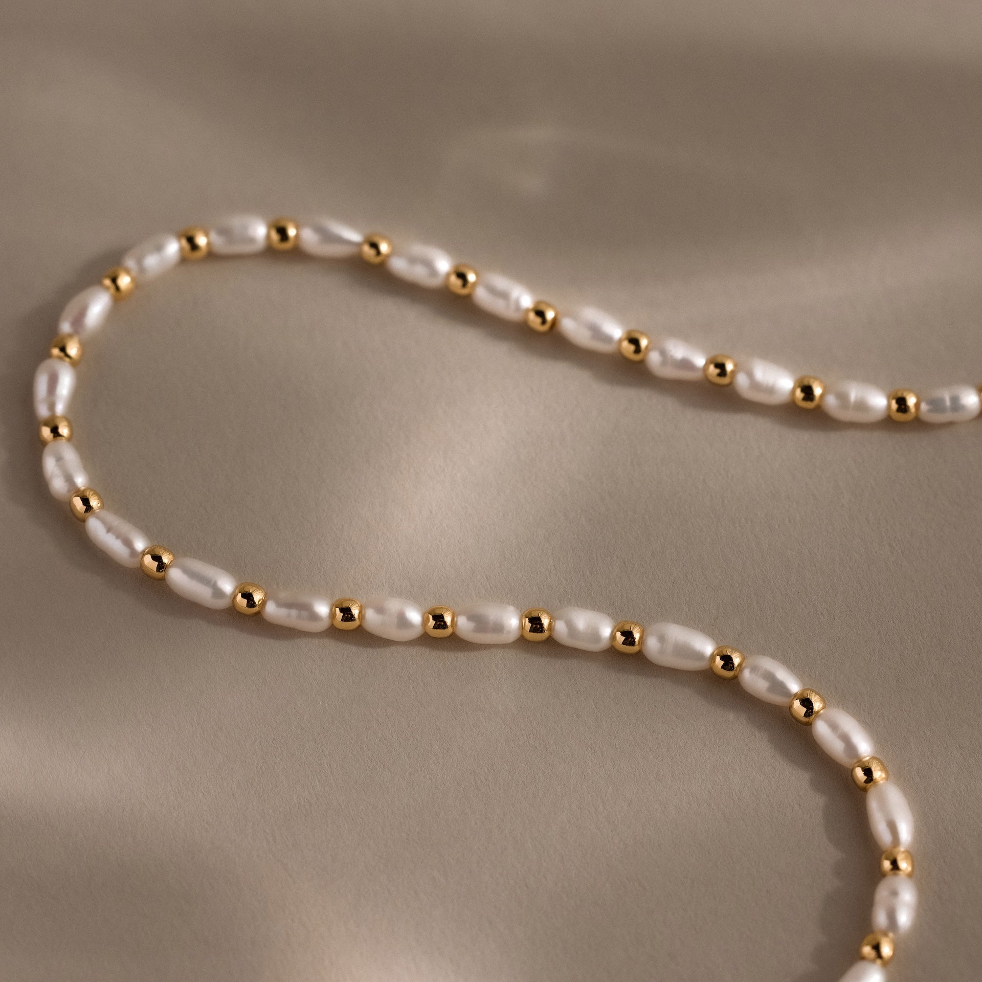 The Beaded Pearl Necklace in 18K Gold features freshwater pearls and polished gold beads, with alternating white pearls and gold accents elegantly arranged on a beige surface.