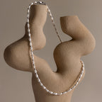 The Beaded Pearl Necklace in White Gold is displayed on an abstract beige sculptural stand, showcasing polished freshwater pearls against a neutral background.