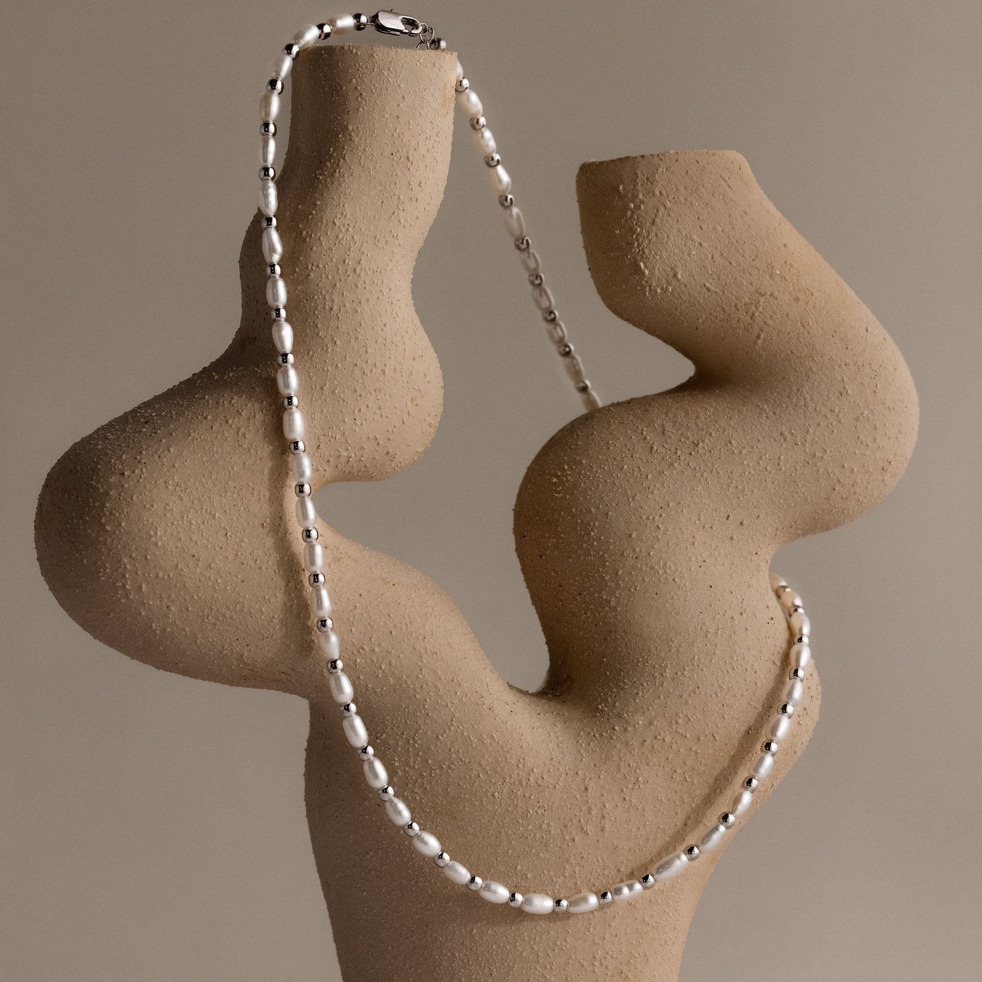 The Beaded Pearl Necklace in White Gold is displayed on an abstract beige sculptural stand, showcasing polished freshwater pearls against a neutral background.