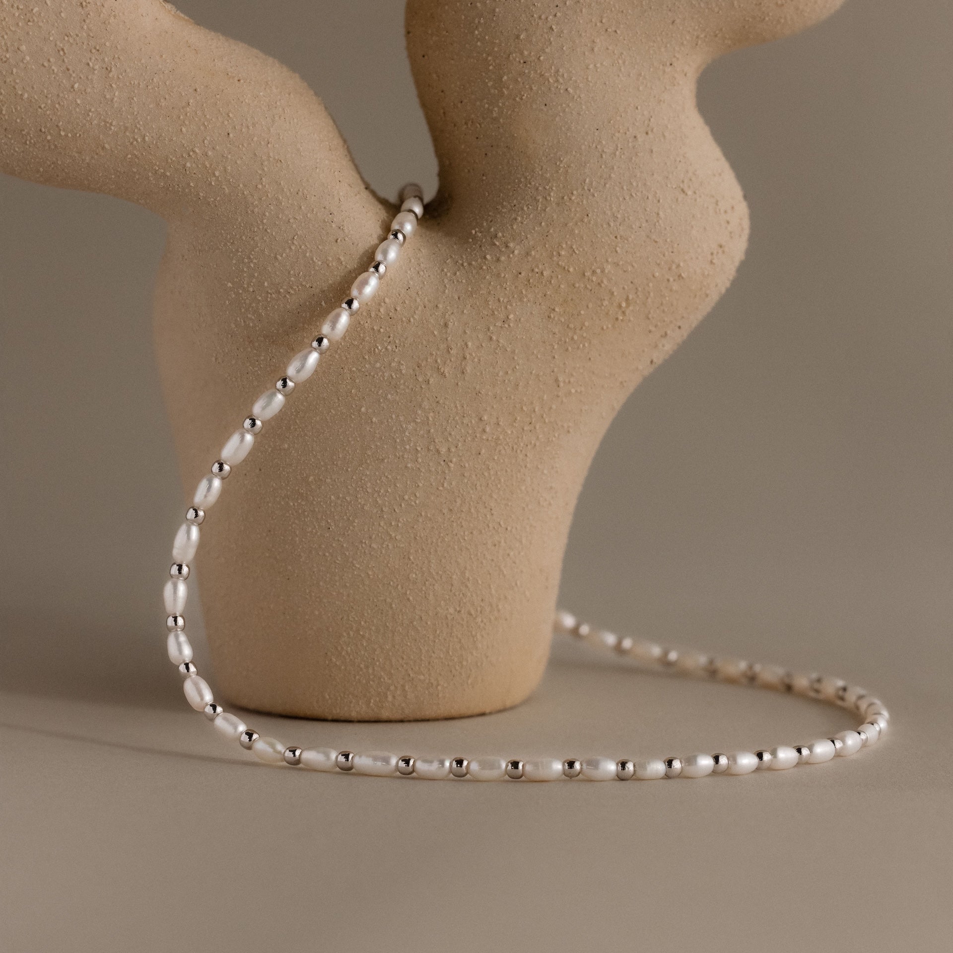 The Beaded Pearl Necklace, featuring delicate polished beads, is draped over a textured beige ceramic vase with a neutral background.