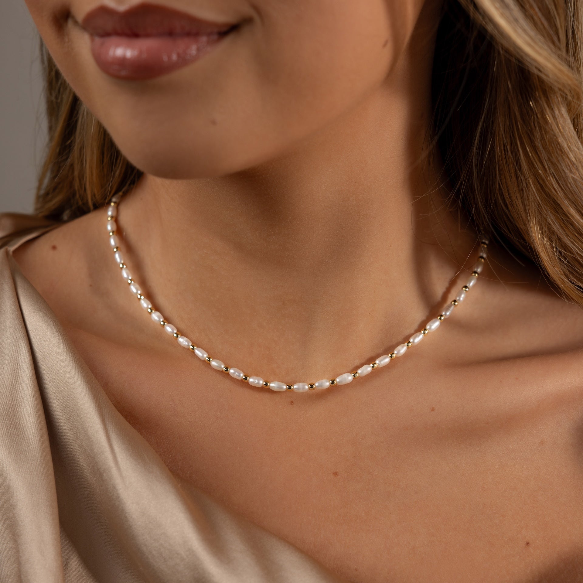 A woman wears the Beaded Pearl Necklace in 18K Gold with a beige satin top, highlighting her neck and part of her face.