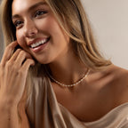 Smiling woman with long hair wearing a beige off-shoulder top and a Beaded Pearl Necklace in 18K Gold adorned with delicate polished beads.