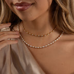 A woman wears a Beaded Pearl Necklace with delicate polished beads layered over a gold chain, paired with a beige satin top.