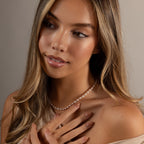 A woman with long, wavy hair wears the Beaded Pearl Necklace in White Gold and touches her chest with manicured hands.