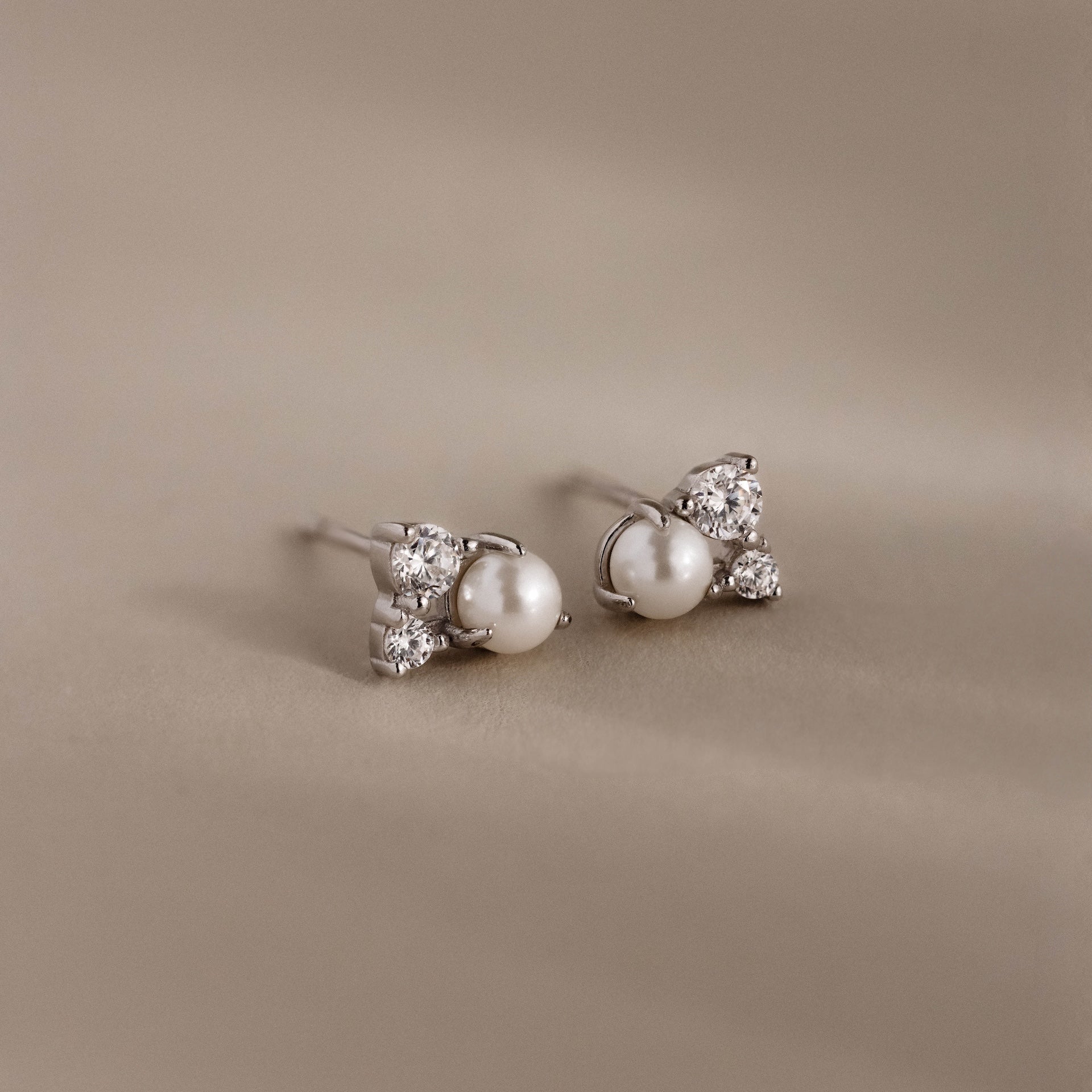 The Pearl Diamond Cluster Studs in Sterling Silver feature three small diamonds accenting pearl studs, displayed on a soft beige background.