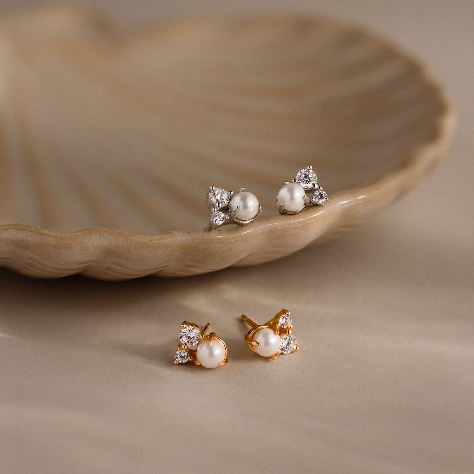 The Pearl Diamond Cluster Studs, available in gold and silver, showcase CZ diamond accents and pearls, elegantly displayed on and near a shell dish.