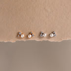 Two pairs of Pearl Diamond Cluster Studs, featuring pearls, CZ diamond accents, and crystals—one in gold and one in silver—are displayed on a textured surface.