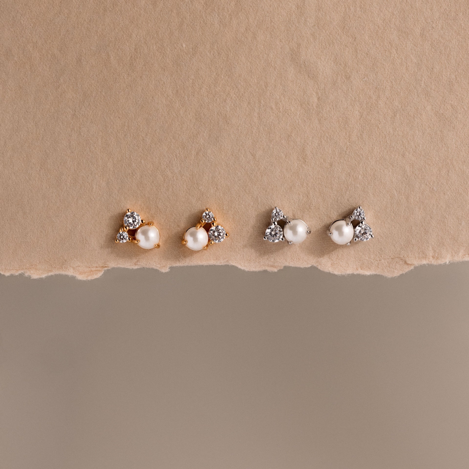 Two pairs of Pearl Diamond Cluster Studs, featuring pearls, CZ diamond accents, and crystals—one in gold and one in silver—are displayed on a textured surface.