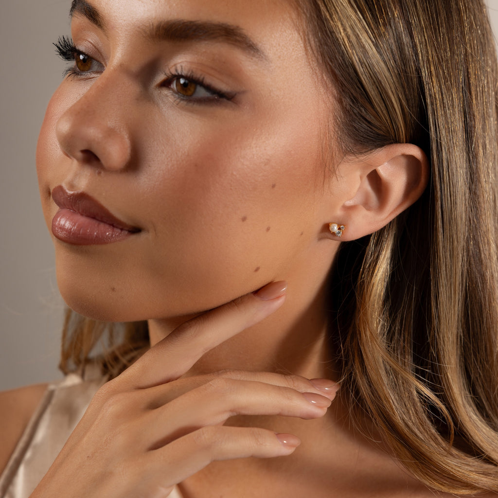 A woman with light brown hair, wearing subtle makeup, touches her chin while showcasing the Pearl Diamond Cluster Studs in 18K Gold—perfect for adding everyday elegance.